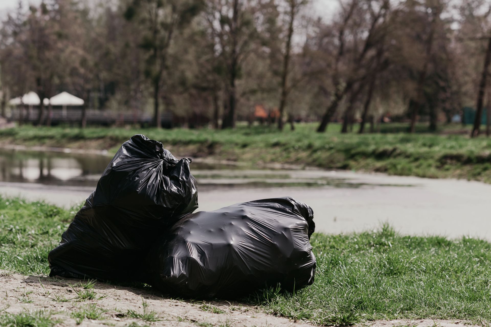 Two black garbage bags on grass by a riverside in a park.